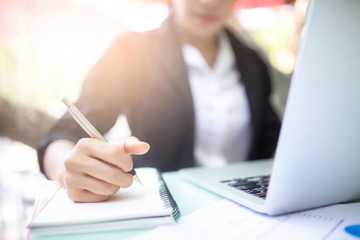 A woman sits at a laptop with a notepad